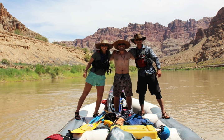 three people stand on a raft and pose for a photo. the raft is floating on water in a canyon.